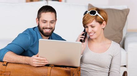 Paar plant eine Reise am Laptop und telefoniert, Koffer im Vordergrund