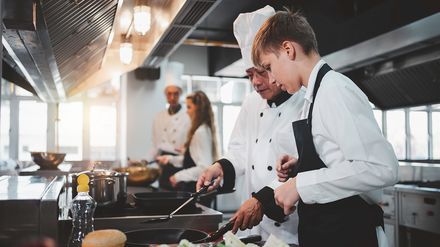 Koch erklärt Auszubildenden in einer professionellen Küche Zubereitungsschritte