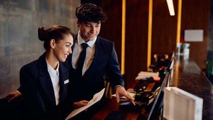 Zwei gut gelaunte Hotelmitarbeiter arbeiten gemeinsam am Empfang eines Hotels und schauen auf einen Computerbildschirm