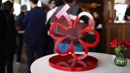 Rote Michelin-Auszeichnung in Form einer stilisierten Blüte auf einem Tisch vor unscharfen Gästen bei einer Veranstaltung.