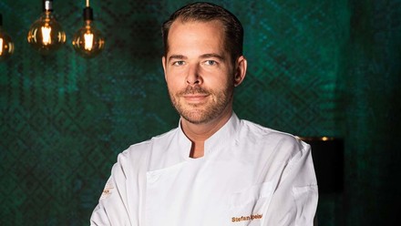 Chefkoch Stefan Speiser sitzt in Kochjacke vor einer dunkelgrünen Wand