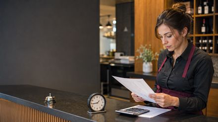 Gastronomie-Mitarbeiterin prüft an einer Restauranttheke einen Dienstplan; neben ihr stehen eine Uhr und ein Taschenrechner.