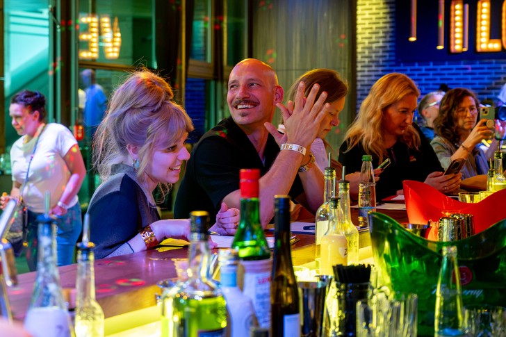 Mehrere Gäste stehen und sitzen an einer Bar, ein Mann in der Bildmitte klatscht.