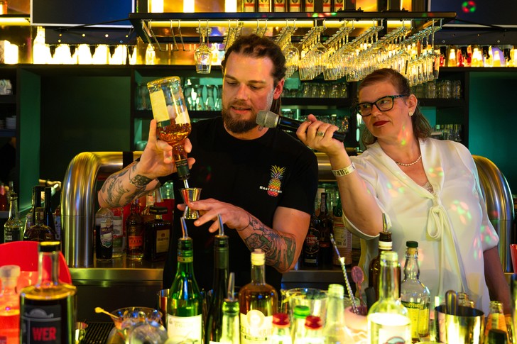 Ein Bartender gießt hinter der Bar eine Flüssigkeit in einen Messbecher, während eine Frau ihm ein Mikrofon hinhält.