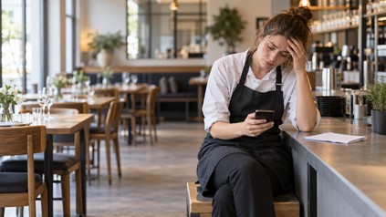 Ein Mitarbeiter im Restaurant sitzt erschöpft an der Theke und informiert den Arbeitgeber per Smartphone über seinen Ausfall.