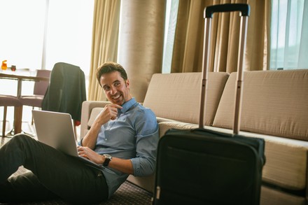 Ein Mann sitzt grinsend vor einer Couch auf dem Boden mit seinem Laptop in einem Hotelzimmer