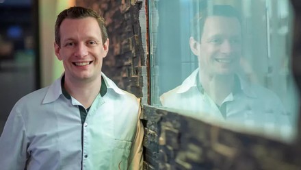 Thomas Schanz mit weißem Hemd lehnt an einer mit Schiefer vertäfelten Wand. Sein Profil spiegelt sich in einer in der Wand eingefassten Milchglasscheibe