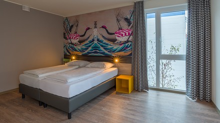Blick in eines der Doppelzimmer des Bento Inn Munich Messe mit einem Doppelbett, einem gelben Nachttisch und bunten Wänden