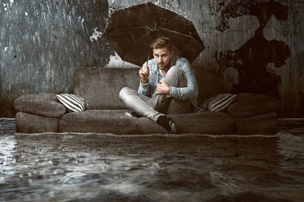 Mann unter Regenschirm auf Sofa in überfluteter Wohnung
