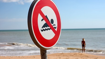 Hotels, die mit direkter Strandlage beworben werden, müssen Urlaubern auch das Schwimmen im Meer vor Ort ermöglichen, wie jetzt das Amtsgericht Hannover urteilte. (Foto: ©Stephane Bonnel/stock.adobe.com)
Strand mit einem „Schwimmen verboten“-Schild, im Hintergrund steht eine Person