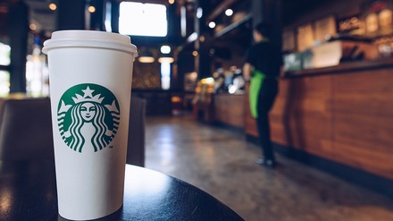 Großer weißer Pappbecher mit grünem Logo von Starbucks im Vordergrund, dahinter verschwommen die Umrisse einer Starbucks Stores
