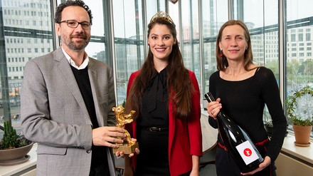 Trafen bei der Auswahlprobe der Weine gemeinsam die Entscheidungen (v.l.n.r.): Künstlerischer Leiter der Berlinale Carlo Chatrian, die Deutsche Weinkönigin Angelina Vogt und Berlinale-Geschäftsführerin Mariette Rissenbeek. (Foto: ©Alex Janetzko)
Zwei Frauen und ein Mann stehen vor einer Fensterfront und halten eine große Weinflasche und den Goldenen Bär, den Hauptpreis der Berlinale, in der Hand