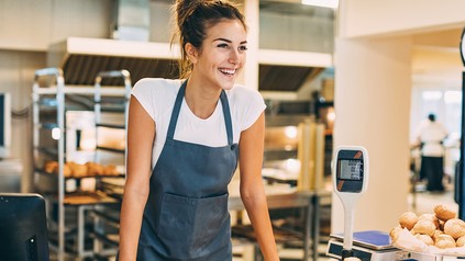 Junge, brünette mit weißem Shirt und blauer Schürze steht lächelnd neben der Lebensmittelwaage in einer Bäckerei