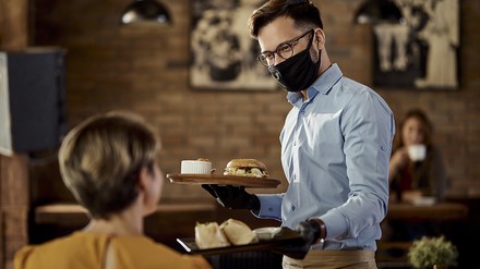Freundlicher Kellner mit Mund-Nasen-Maske bedient eine Frau im Restaurant