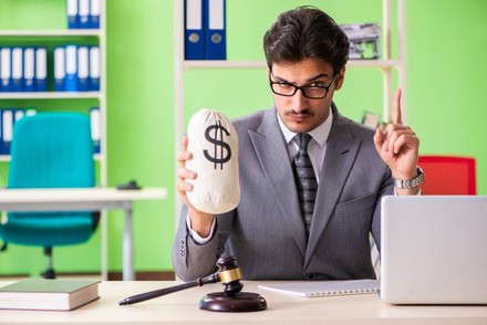 Ein Beamter vor einem Computer hält einen Sack Geld in der Hand und hebt mahnend den Finger