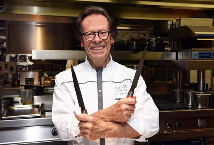 Spitzenkoch Dieter Müller steht mit zwei Messern lächelnd in einer Küche