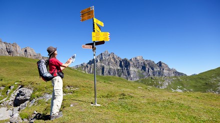 Frau in Wanderkleidung mit einem Wanderrucksack steht vor einem Schilderbaum und schaut nach dem Weg