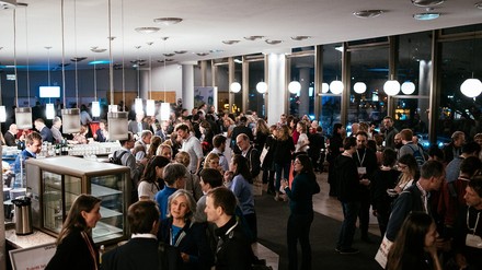 Viele Menschen stehen in einem Foyer an Bistrotischen und unterhalten sich
