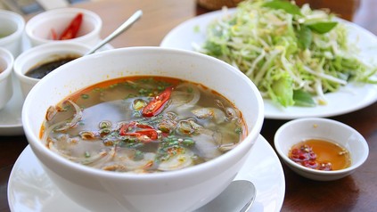 Eine Schüssel mit Nudelsuppe, mit Flesich und Gemüse zubereitet, im Hintergrund ein grüner Salat