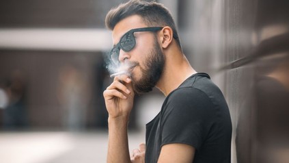 Mann mit Sonnenbrille lehnt an einer Wand und raucht eine Zigarette
