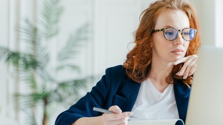 Frau mit Brille und Stift in der Hand vor dem Laptop