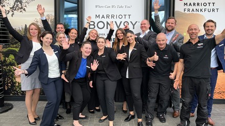 Mitarbeiter des Frankfurt Marriott Hotels sind gut gelaunt – denn „Work-Live-Balance“ und „Take-Care“ werden hier großgeschrieben. (Foto: © Frankfurt Marriott Hotel) Gruppenfoto der Mitarbeiter des Marriott Bonvoy in Frankfurt, die mit ausladenden Gesten vor der Fassade des Hotels posieren