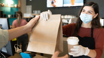 Junge Barista mit Mund-Nasen-Maske und Handschuhen gibt eine Papiertüte mit Essen und einen Kaffee an einen Kunden aus