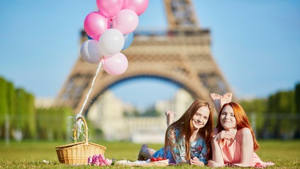 Zwei junge Frauen liegen mit einer Decke und einem Picknickkorn auf der Wiese vor dem Eifelturm
