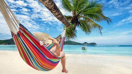 Eine Frau liegt mit einem Sonnenhut entspannt an einer Hängematte an einer Palme am Strand