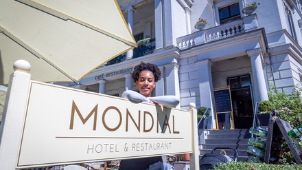 Joy Ewulu in der Rolle der Lara Hildebrand steht in einer Pause bei Dreharbeiten für die neue ZDF-Serie „Hotel Mondial“ hinter dem Hotelschild. (Foto: © picture alliance/dpa | Jens Büttner)
Joy Ewulu vor dem Hotelschild des „Hotel Mondial“