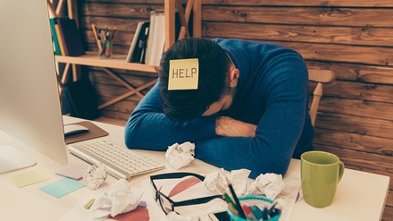 Wenn einen die Müdigkeit im Job überfällt, dann braucht es schnell wirkende Tipps. (Foto: ©deagreez/stock.adobe.com)
Müder junger Mann hat die Arme erschöpft auf den Schreibtisch gelegt und seinen Kopf darauf gestützt, auf seinem Kopf klebt ein gelbes Post-it mit der Aufschrift Help