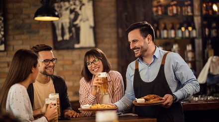 Der BVMW schlägt einen deutschlandweiten Aktionsplan vor, um wieder mehr Menschen für das Arbeiten in der Tourismusbranche zu begeistern. (Foto: © Drazen/stock.adobe.com)
Gruppe wird im Wirtshaus von lächelndem Kellner bedient
