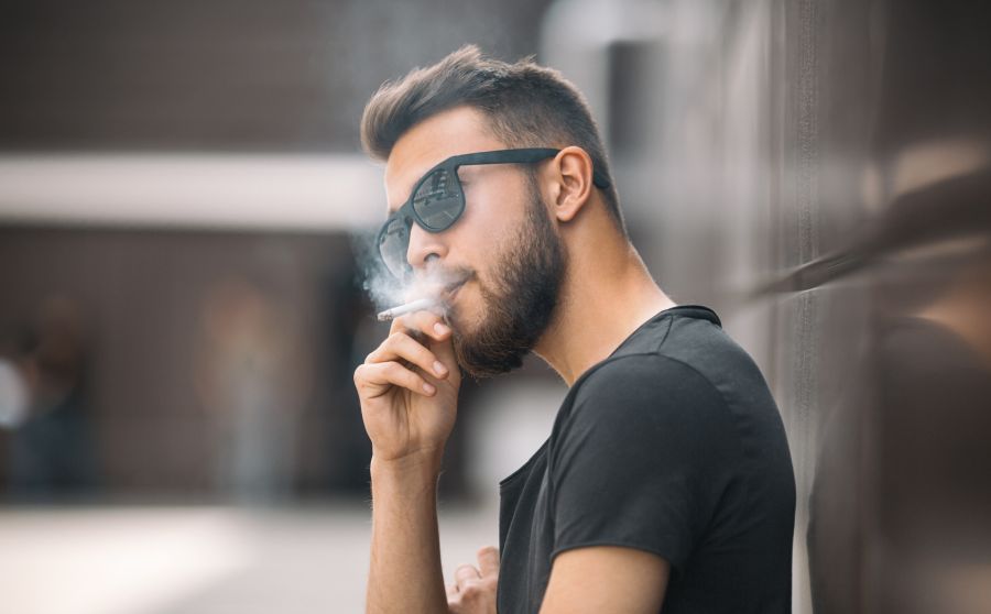 Mann mit Sonnenbrille lehnt an einer Wand und raucht eine Zigarette